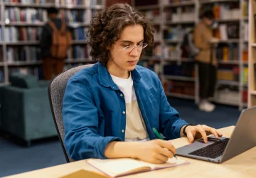 Estudante pesquisando em sites de divulgação científica no notebook durante estudo na biblioteca.