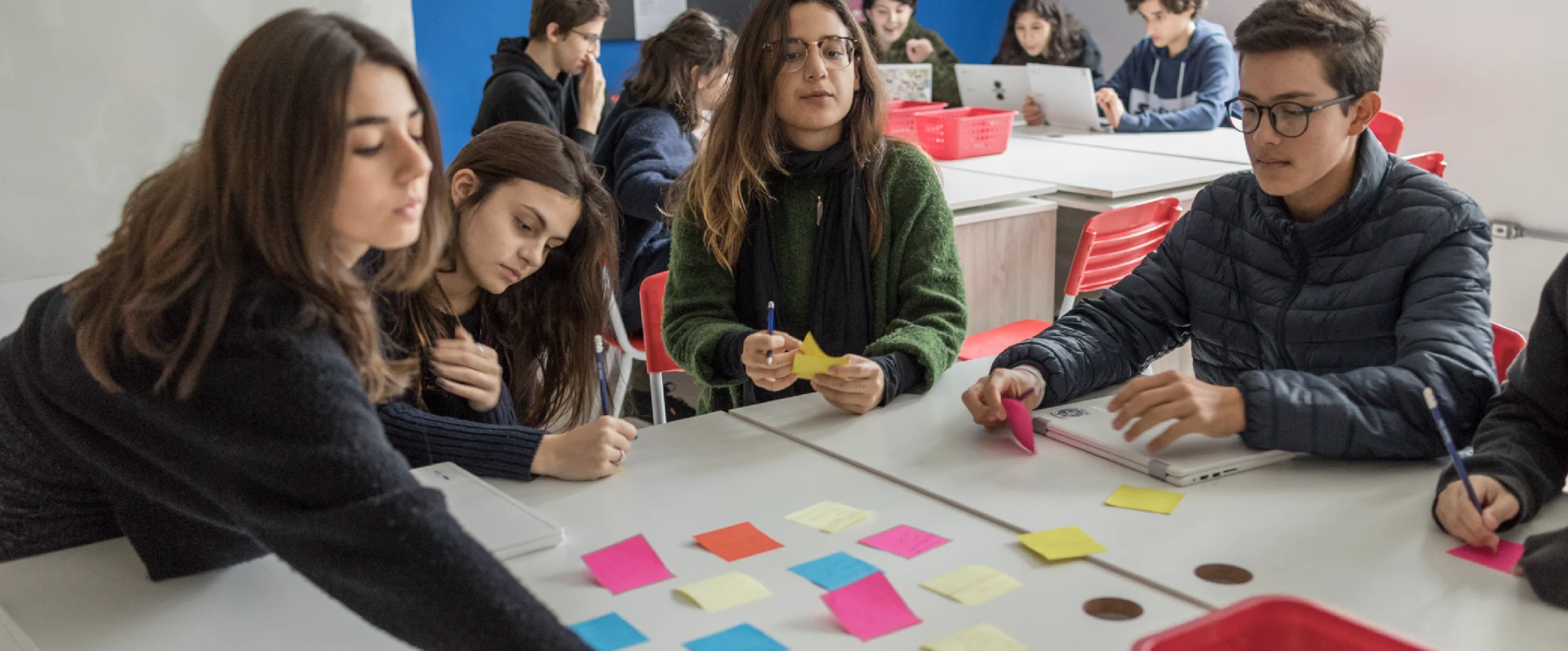 Estudantes trabalham em grupo com post-its e notebook em uma atividade de rotação por estações.