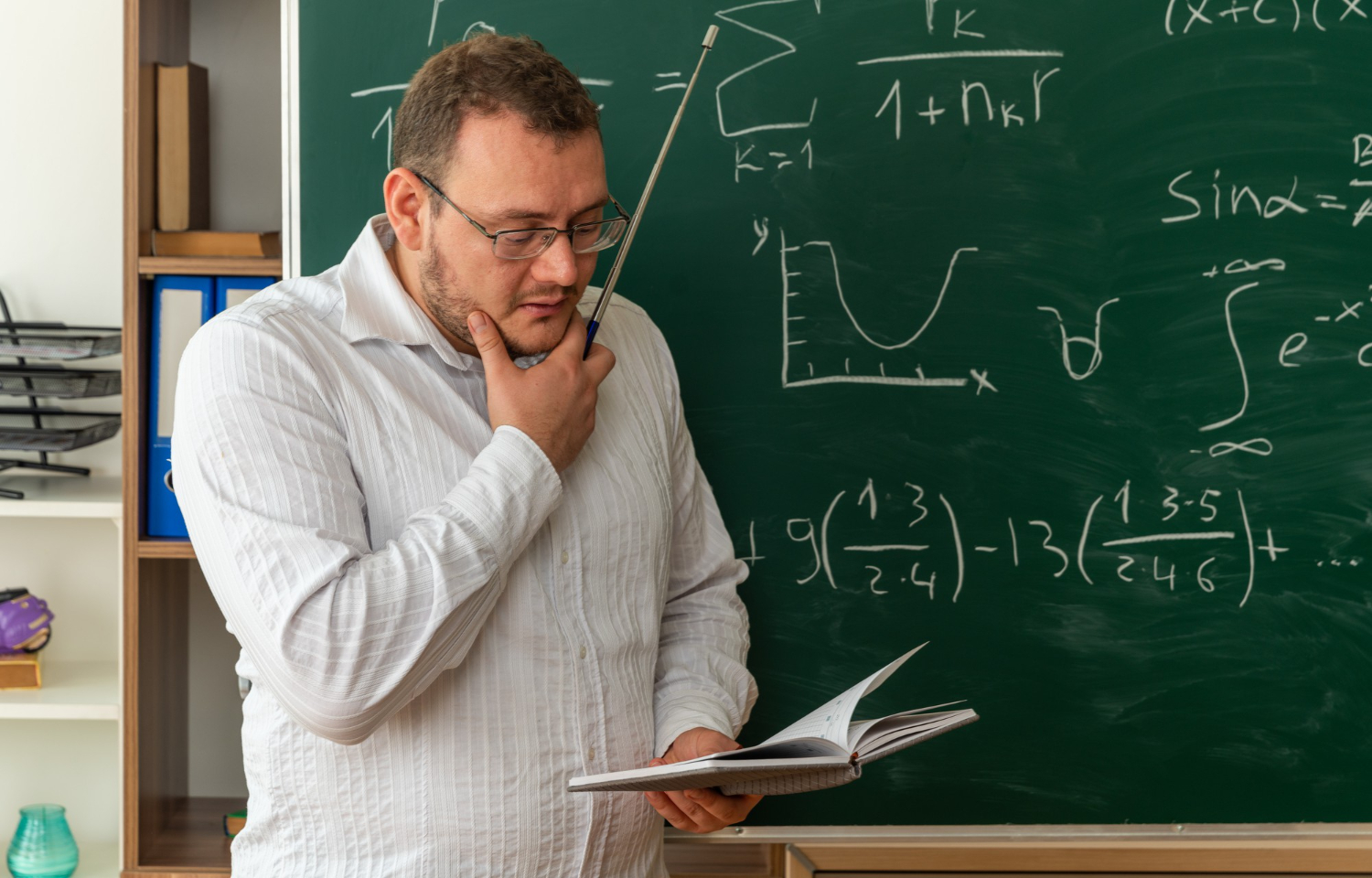 Professor consulta livro diante da lousa, representando objetivos de aprendizagem BNCC