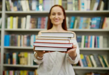 Professora mostra pilha de livros de material didático para o ensino fundamental em biblioteca escolar.