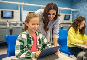 Professora orientando aluna no uso de tablet durante atividade de formação docente com tecnologia educacional.
